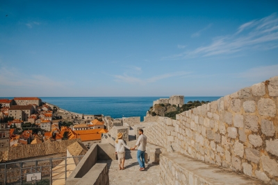Wide panoramic photo taken at Dubrovnik City Walls, couple walking on a photo shoot in the morning. Old town red roofs, st. Lawrence fortress and blue sea and sky in the background